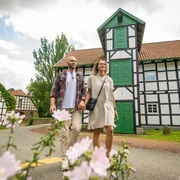 Gutswassermühle Hudenbeck Ein Paar spaziert vor einer Fachwerk-Gutswassermühle, mit blühenden Blumen im Vordergrund.