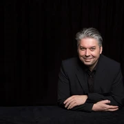 BlackTableMagic.jpg Mann mit Anzug stützt sich auf seine Ellbogen und lächelt in die Kamera.