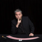 Mann hält sich seinen Zeigefinder vor den Mund und stützt sich auf einen Tisch, auf dem Spielkarten halbkreisförmig ausgebreitet sind.