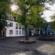 Kellerplatz in Beverungen Der Kellerplatz in Beverungen zeigt ein weißes Rathaus mit Fachwerkgiebel. Ein Brunnen mit St.-Michaelsfigur steht im Mittelpunkt, umgeben von schattenspendenden Bäumen, die dem Platz eine einladende Atmosphäre verleihen. Diese Szene betont die historische und zugleich lebendige Präsenz des Ortes.