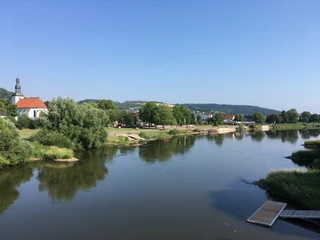 Weserpromenade in Beverungen Das Foto zeigt den Verlauf des Weserflusses mit Uferbereich. Auf der linken Seite befindet sich eine weiße Kirche, umgeben von Bäumen, und im Hintergrund erstreckt sich ein bergiges Hinterland.