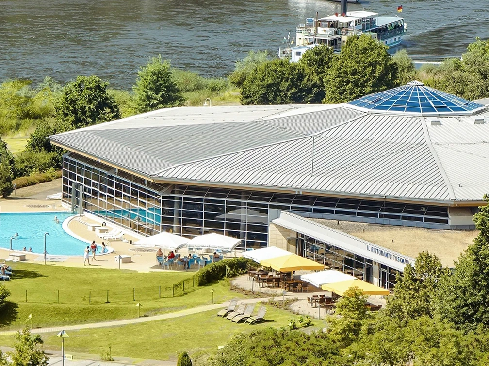 Il Ristorante Toskana Modernes Gebäude mit großem Glasdach, umgeben von Bäumen, mit einem Außenpool und einer Terrasse mit Sonnenschirmen; im Hintergrund ein Fluss.