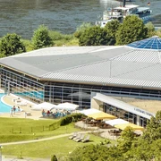 Il Ristorante Toskana Modernes Gebäude mit großem Glasdach, umgeben von Bäumen, mit einem Außenpool und einer Terrasse mit Sonnenschirmen; im Hintergrund ein Fluss.