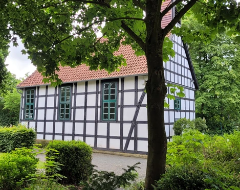 Fachwerkgebäude inmitten von Bäumen, mit weißen Wänden und schwarzen Balken; rote Ziegeldach.