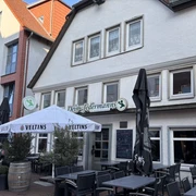 Jedermann_3.jpg Außenansicht eines Restaurants mit Terrasse, Holzmöbeln und Sonnenschirmen unter blauem Himmel.