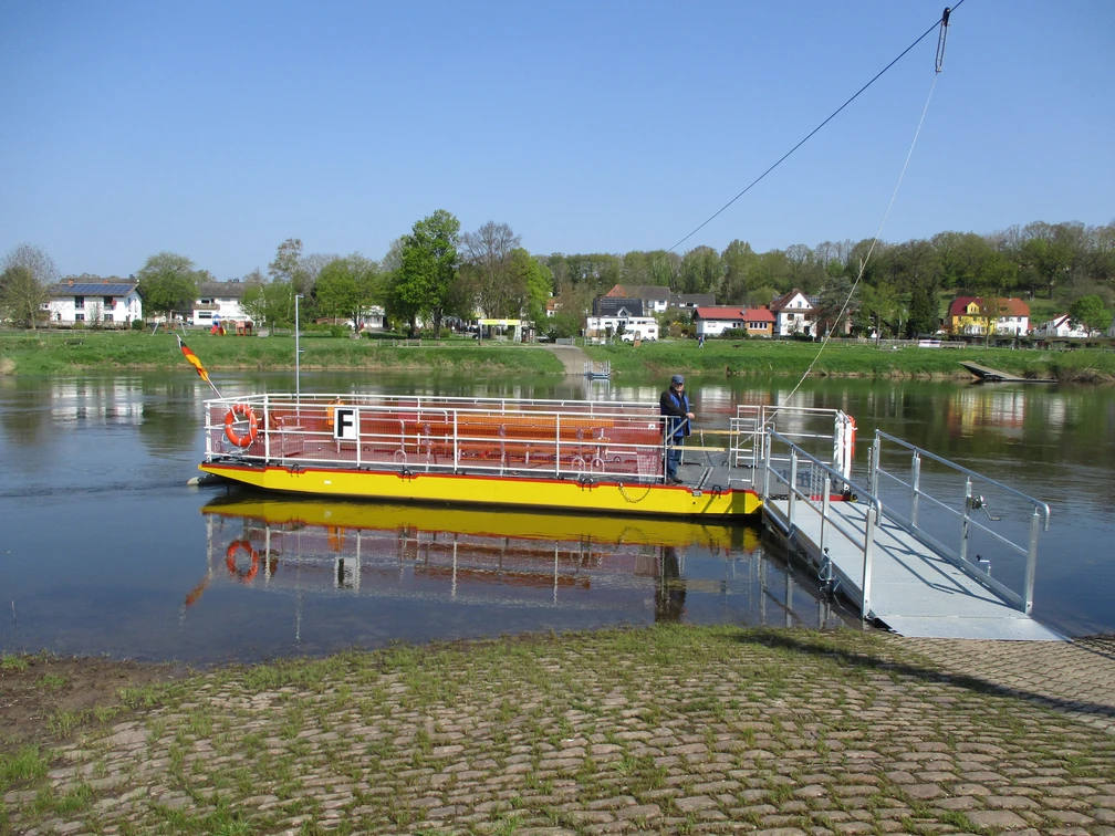 Dreiländereckfähre Herstelle/Würgassen Eine gelbe Seilfähre überquert die Weser, im Hintergrund eine Kulisse aus Bäumen und Häusern.