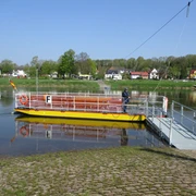 Dreiländereckfähre Herstelle/Würgassen Eine gelbe Seilfähre überquert die Weser, im Hintergrund eine Kulisse aus Bäumen und Häusern.