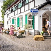 Außenbereich Tante Tina Das Bild zeigt den Außenbereich von Tante Tina mit Gästen, die auf einer Kopfsteinpflasterstraße sitzen.