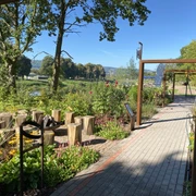 schoepfungsgarten_c_team_schoepfungsgarten (3).jpeg Grünanlage mit gepflastertem Weg, Pergola und Feuerschale mit Holzstamm Sitzhockern