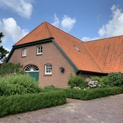 Hof Kleiberg Ein rotes Backsteinhaus mit spitzem Dach, umgeben von grünen Hecken und bunten Blumen, vor blauem Himmel.