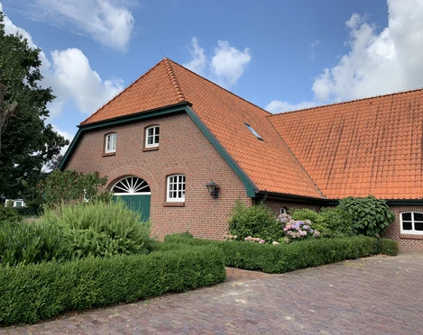 Hof Kleiberg Ein rotes Backsteinhaus mit spitzem Dach, umgeben von grünen Hecken und bunten Blumen, vor blauem Himmel.