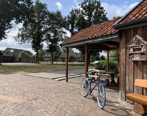 Hof Kleiberg Ein Hof mit Holzhaus, Fahrrad, grünem Baum und Bank auf gepflastertem Platz bei blauem Himmel.
