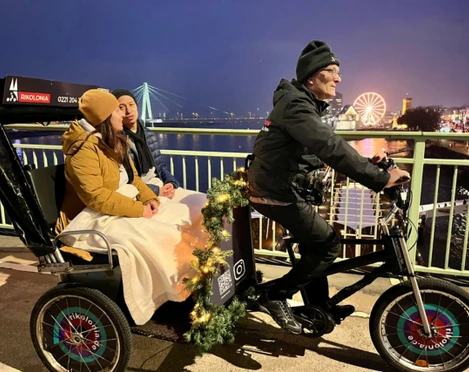 Rikolonia Köln Wintertouren II.png Rikscha fährt über eine Brücke über den Rhein und transportiert zwei PersonenRickshaw crosses a bridge over the Rhine and transports two people