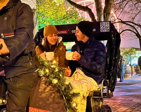 Rikolonia Köln Glühweinfahrt.png Zwei Personen sitzen am Abend hinten in einer Rikscha und trinken GlühweinTwo people sit in the back of a rickshaw in the evening and drink mulled wine