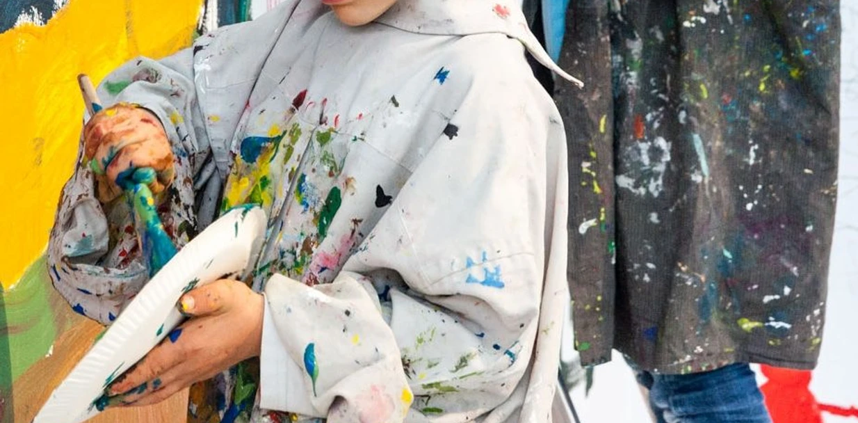 Sonntagsmalerei_LudwigForum-Foto-Carl-Brunn.jpg Ein Kind malt konzentriert an einer Leinwand, bekleidet mit einem übergroßen, buntspritzigen Malkittel.