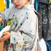 Sonntagsmalerei_LudwigForum-Foto-Carl-Brunn.jpg Ein Kind malt konzentriert an einer Leinwand, bekleidet mit einem übergroßen, buntspritzigen Malkittel.