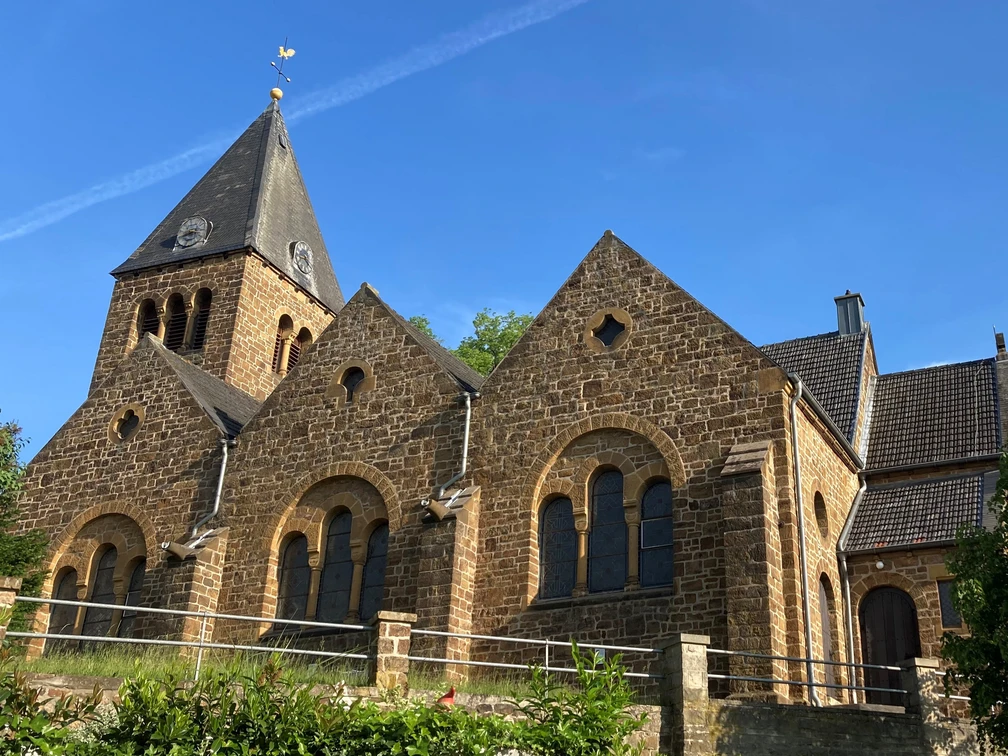 Braune Backsteinkirche mit hohen Giebeln und Kirchturm vor blauem Himmel, umgeben von Bäumen.