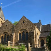 Braune Backsteinkirche mit hohen Giebeln und Kirchturm vor blauem Himmel, umgeben von Bäumen.