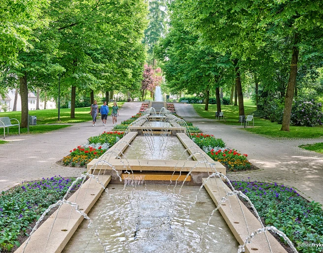 Brunnen im Kneipp-Kurpark Bad Camberg In der Mitte ist ein Kaskadenbrunnen zu sehen, links und rechts davon laufen jeweils zwei Personen.