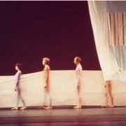Stage design for Cunningham's choreography Travelogue Vor einem dunklen Bühnenhintergrund stehen in einer Reihe mehrere Menschen eng zusammen, eingehüllt in durchscheinende, farbig leuchtende Stoffbahnen, die gemeinsam ein ästhetisches Bild voller Anmut und Bewegung formen.Against a dark stage backdrop, several people stand close together in a row, wrapped in translucent, brightly colored lengths of fabric, which together form an aesthetic image full of grace and movement.