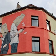 Mural Auf dem Bild ist ein großes Wandgemälde eines Gebäudes an der Ecke einer Straße zu sehen. Zwei Figuren stehen Rücken an Rücken, beide mit Tüten über dem Kopf und Handys in der Hand. Ein Pfeil fliegt von hinten auf die Figuren zu, als ob er von Amor, der links im Bild erscheint, abgeschossen wurde. Die Farben sind hauptsächlich beige und rot, mit schwarzen Fensterumrandungen, die das Gemälde unterbrechen.The picture shows a large mural of a building on the corner of a street. Two figures are standing back to back, both with bags over their heads and cell phones in their hands. An arrow flies towards the figures from behind, as if it has been shot by Cupid, who appears on the left of the picture. The colors are mainly beige and red, with black window borders interrupting the painting.