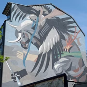 Mural Ein dynamischer Stierkampf zwischen Stier und abstraktem Wesen, in kräftigen Farben an einer Hauswand, von grünen Bäumen eingerahmt.A dynamic bullfight between a bull and an abstract creature, in strong colors on a house wall, framed by green trees.