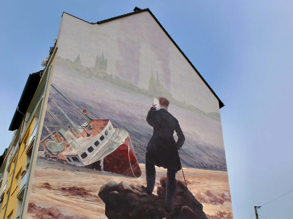 Mural Das Bild zeigt ein Wandgemälde an einem Haus, auf dem ein stilisierter Wanderer steht. Der Wanderer blickt auf ein gestrandetes Schiff vor einer historischen Städtekulisse.The picture shows a mural on a house with a stylized wanderer standing on it. The wanderer is looking at a stranded ship in front of a historic city backdrop.