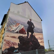 Mural Das Bild zeigt ein Wandgemälde an einem Haus, auf dem ein stilisierter Wanderer steht. Der Wanderer blickt auf ein gestrandetes Schiff vor einer historischen Städtekulisse.The picture shows a mural on a house with a stylized wanderer standing on it. The wanderer is looking at a stranded ship in front of a historic city backdrop.