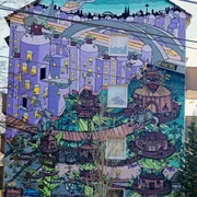 Mural Lebendige Street Art in Köln: Ein Hochhaus mit farbenfrohem Wandgemälde, das eine fantasievolle Stadtlandschaft zeigt, in der überdimensionale Spielfiguren und architektonische Elemente in surrealer Anordnung zu sehen sind. Auffallend sind die dominanten Blau- und Lilatöne sowie die verspielten, märchenhaften Details.Vibrant street art in Cologne: A high-rise building showcasing a colorful mural depicting an imaginative cityscape, featuring oversized game pieces and architectural elements arranged in a surreal manner. Prominent blue and purple tones, along with playful, fairy-tale details, stand out distinctly.