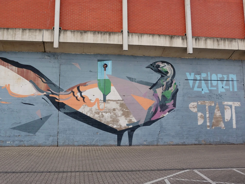 Mural Auf dem Bild ist ein farbenfrohes Graffiti an einer Betonmauer zu sehen. Es zeigt eine künstlerische Darstellung eines Vogels, der in geometrischen Formen und kräftigen Farben gestaltet ist. Oberhalb des Bildes ist eine Backsteinwand zu erkennen, die das Motiv rahmt. The picture shows a colorful graffiti on a concrete wall. It shows an artistic depiction of a bird designed in geometric shapes and bold colors. A brick wall can be seen above the picture, framing the motif.