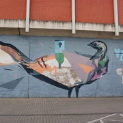 Mural Auf dem Bild ist ein farbenfrohes Graffiti an einer Betonmauer zu sehen. Es zeigt eine künstlerische Darstellung eines Vogels, der in geometrischen Formen und kräftigen Farben gestaltet ist. Oberhalb des Bildes ist eine Backsteinwand zu erkennen, die das Motiv rahmt. The picture shows a colorful graffiti on a concrete wall. It shows an artistic depiction of a bird designed in geometric shapes and bold colors. A brick wall can be seen above the picture, framing the motif.