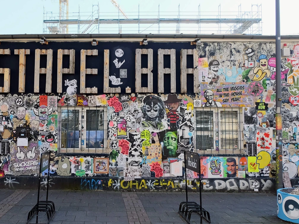 Mural Eine bunte Street-Art-Wand zeigt Graffiti und Sticker in lebhaften Farben, flankiert von Baugerüsten.A colorful street art wall displays graffiti and stickers in vibrant colors, flanked by scaffolding.