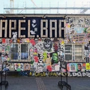 Mural Eine bunte Street-Art-Wand zeigt Graffiti und Sticker in lebhaften Farben, flankiert von Baugerüsten.A colorful street art wall displays graffiti and stickers in vibrant colors, flanked by scaffolding.