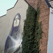 Mural Das Kunstwerk zeigt eine beeindruckende, großflächige Wandmalerei in Köln. Das Bild zeigt eine Madonna mit geschlossenen Augen, goldene Akzente betonen ihr Gesicht. Die Szene wird von üppigem Efeu flankiert, das an der Backsteinmauer hochwächst und einen harmonischen Kontrast zur urbanen Umgebung bildet. The artwork shows an impressive, large-scale mural in Cologne. The picture shows a Madonna with closed eyes, golden accents emphasize her face. The scene is flanked by lush ivy growing up the brick wall, creating a harmonious contrast to the urban surroundings.