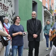 Vringsveedels tour with Wachter Loup Auf dem Bild ist eine Gruppe von Menschen unterschiedlicher Altersklassen zu sehen, die auf einer Straße in Köln steht. Zwei Männer haben Gitarren in der Hand, während ein weiterer Mann im Anzug in der Mitte steht. Einige Personen hören interessiert zu, während sie vor einer Wand mit Graffiti stehen. The picture shows a group of people of different ages standing on a street in Cologne. Two men are holding guitars, while another man in a suit stands in the middle. Some people are listening with interest as they stand in front of a wall with graffiti.