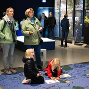 Weihnachtlicher Familiennachmittag_nbz-Alfsee.jpg Drei Erwachsene beobachten Kinder, die auf einem großen, blauen Bodenplan in einem Ausstellungsraum spielen.