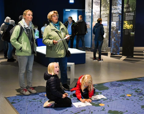Weihnachtlicher Familiennachmittag_nbz-Alfsee.jpg Drei Erwachsene beobachten Kinder, die auf einem großen, blauen Bodenplan in einem Ausstellungsraum spielen.