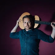 Philipp Godart Ein Mann in dunkelblauem Hemd hält eine Akustikgitarre lässig quer über seine Schultern.A man in a dark blue shirt holds an acoustic guitar casually across his shoulders.