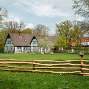 Fachwerkgebäude mit dunklem Dach, umgeben von grüner Wiese, Bäumen und Schafen hinter Holzzaun.