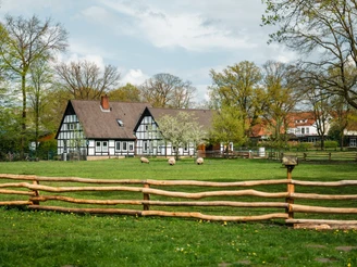 Museumshof Rahden  Fachwerkgebäude mit dunklem Dach, umgeben von grüner Wiese, Bäumen und Schafen hinter Holzzaun.