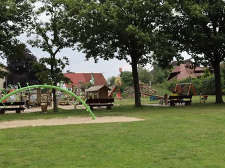 Spielplatz mit Klettergerüst, Schaukeln und Rasenflächen umgeben von großen Bäumen, ideal für Familien.