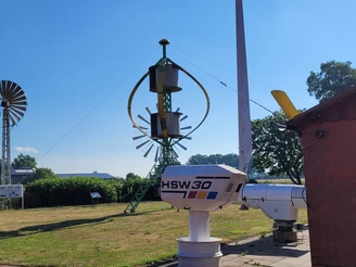 Windkraftmuseum mit alten Windturbinen und technischen Anlagen auf einer Wiese bei blauem Himmel.