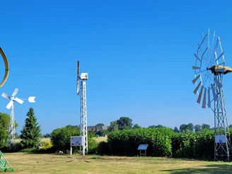 Verschiedene historische Windkraftanlagen auf grüner Wiese, umgeben von Bäumen und blauem Himmel.