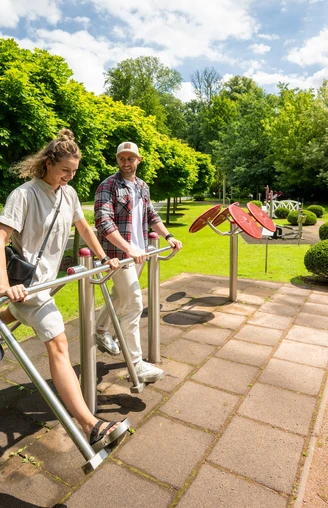 Besucher genießen die Nutzung von Outdoor-Fitnessgeräten inmitten einer grünen Parklandschaft.