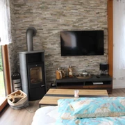 Ein helles Wohnzimmer mit Kaminofen, Flachbildfernseher und großen Fenstern, Blick in den Garten.