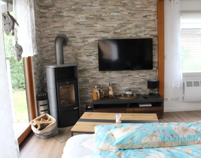 Ferienhaus Max Ein helles Wohnzimmer mit Kaminofen, Flachbildfernseher und großen Fenstern, Blick in den Garten.