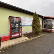 Proviantomat Standort Struppen Landfleischerei Roter Verkaufsautomat mit Lebensmitteln vor einem hellgrünen Gebäude, daneben ein Schild mit Angeboten und eine kleine, konische Tanne.