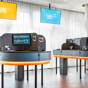 Rezeption im Hey Lou Monheim am Rhein Moderne Rezeption mit zwei Servicepunkten, beleuchtetem Tresen, digitalen Bildschirmen und hellen Vorhängen.