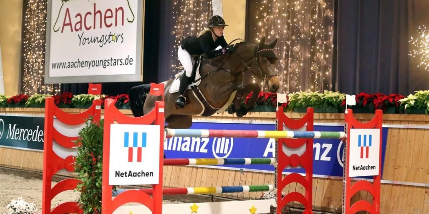 Eine Reiterin springt mit einem Pferd über ein Hindernis in einer weihnachtlich geschmückten Reithalle.A rider jumps over an obstacle with a horse in an indoor riding arena decorated for Christmas.Une cavalière saute un obstacle avec son cheval dans un manège décoré pour Noël.Een ruiter springt met een paard over een hindernis in een overdekte rijhal die versierd is voor Kerstmis.