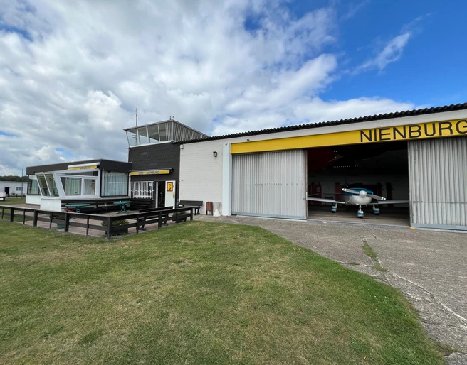 Flugplatz mit Hangars und einer gelben Markierung "Nienburg", Wiese im Vordergrund.